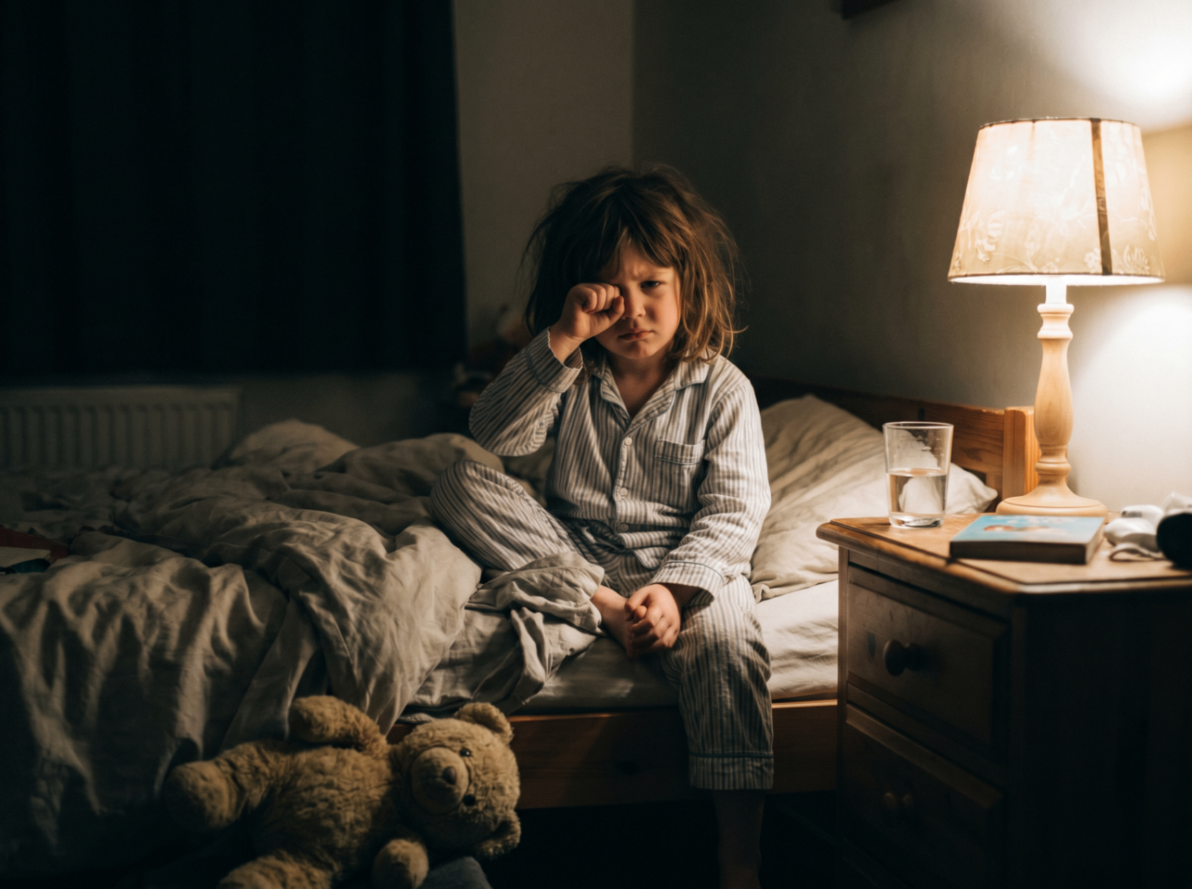 A tired young child sitting on the edge of the bed at night, rubbing her eyes